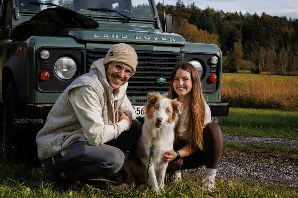 mann mit Mütze, Hund in der Mitte und Frau mit langem haar sitzen vor einen grünen land Rover Defender